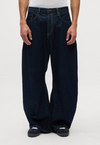 Pantalones vaqueros de pierna ancha en denim oscuro con costuras amarillas en contraste y diseño de cinco bolsillos. La cintura cuenta con un cierre de botón metálico.