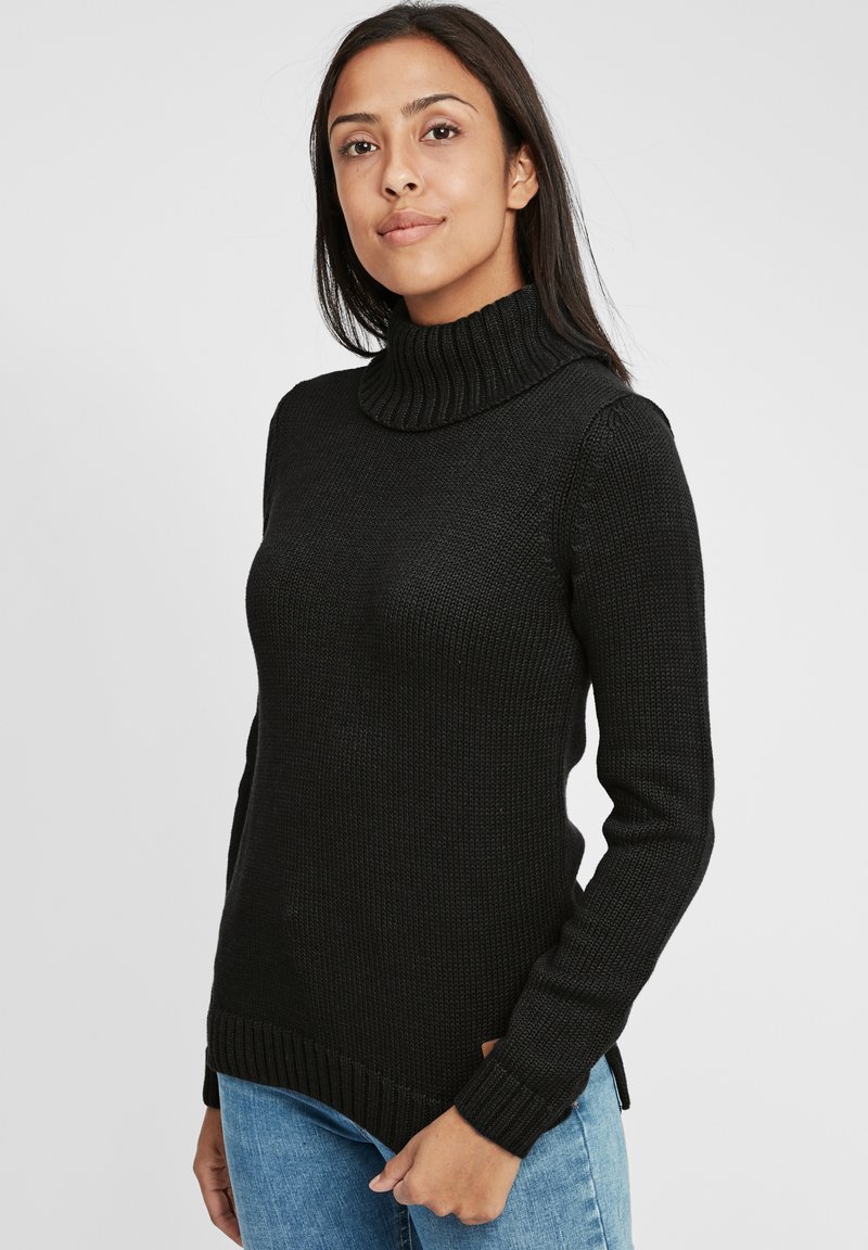 Schwarzer gestrickter Pullover mit hohem Rollkragen, gerippter Textur, langen Ärmeln und seitlichen Schlitzen, kombiniert mit hellblauen Jeans.