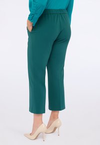 Kobieta w turkusowych, krótkich spodniach z kieszeniami i beżowych szpilkach, stojąca z rękami w kieszeniach na białym tle.