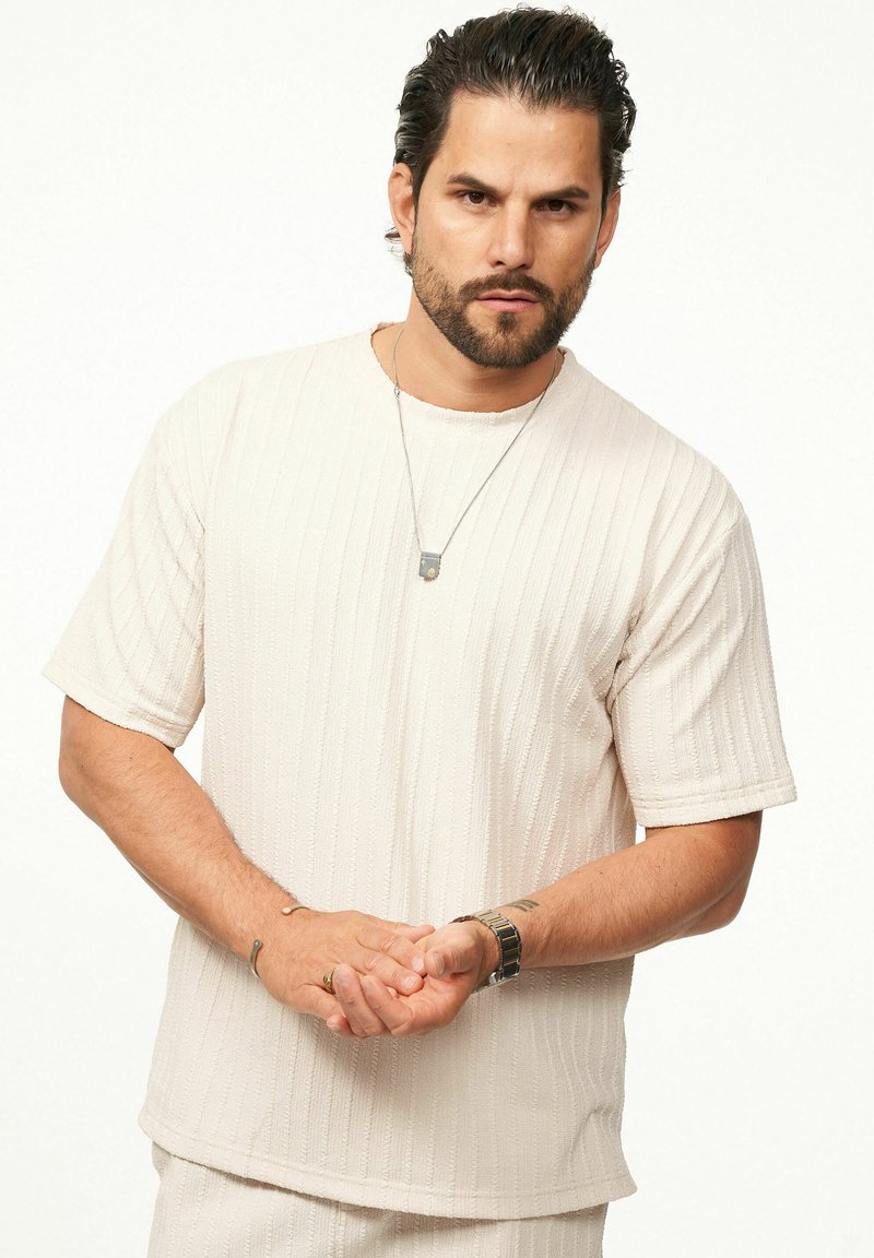 Beige strukturiertes Strick-T-Shirt mit kurzen Ärmeln und einem gerippten Muster. Kombiniert mit silberfarbenem Schmuck und einer Uhr.