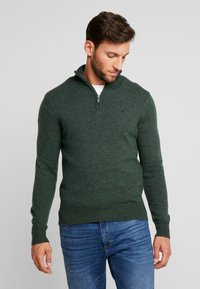 Suéter verde con cremallera y tejido de punto texturizado. Presenta un cuello alto y puños y dobladillo de canalé. Se lleva sobre una camisa blanca, combinado con vaqueros.