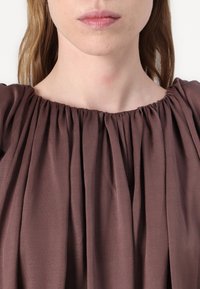 Brown, pleated blouse with a gathered neckline. Soft fabric with a smooth texture, featuring deep folds that create visual interest.