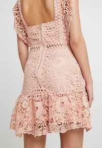 Růžové krajkové šaty s propracovanými květinovými vzory, přiléhavým korzetem se zipovým uzávěrem a rozšířeným volánkovým lemem.