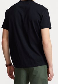 Persoon in een eenvoudige zwarte korte mouw shirt en olijfgroene broek, gezien van achteren, met een bruine horloge om de linker pols.