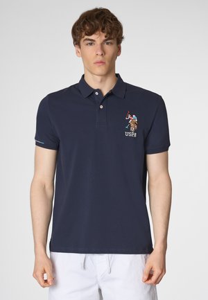 Un giovane uomo è in piedi indossando una polo blu navy con un logo USPA ricamato. Lo sfondo è semplice, mettendo in risalto il design della maglietta.