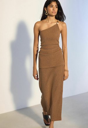 Woman in a brown one-shoulder textured dress with wooden beads on the strap, wearing gold hoop earrings and black heeled sandals.
