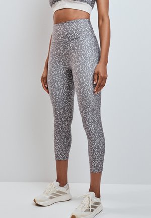 Donna che indossa leggings corti grigi con motivo, reggiseno sportivo bianco e scarpe da corsa bianche, in piedi davanti a uno sfondo semplice.