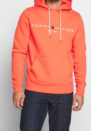 Hoodie orange avec une poche avant, des cordons blancs et un logo qui indique "TOMMY HILFIGER EST. 1985" sur la poitrine. Texture lisse.