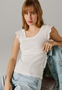 Noor naine, kes kannab õlgadeni juustega valget varrukateta topi silmkoeliste pitsiliste varrukatega ja helesiniseid teksaseid, hoides teksajopet.