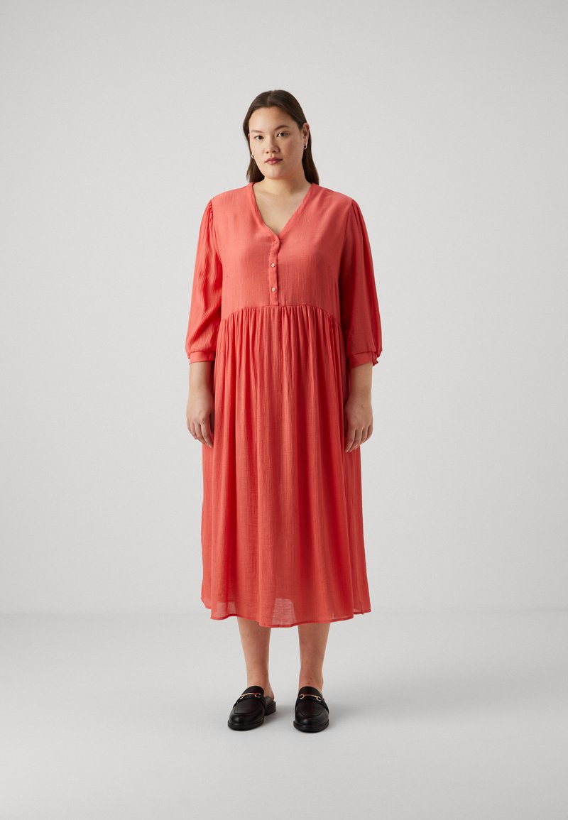 Koraļļu krāsas kleita līdz ceļgaliem, kas izgatavota no viegla auduma, ar V veida kakla izgriezumu, trīs ceturtdaļu piedurknēm un pogu detaļām uz krūšu daļas.