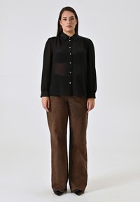 Blusa nera trasparente con colletto e chiusura a bottoni, abbinata a pantaloni larghi marroni. Scarpe con tacco nere completano l'outfit.