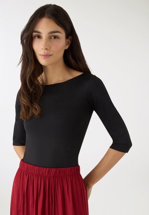 Kobieta o długich ciemnych włosach, ubrana w dopasowaną czarną bluzkę z rękawami do łokcia oraz czerwoną spódnicę z wysokim stanem, stojąca na tle jednolitego tła.