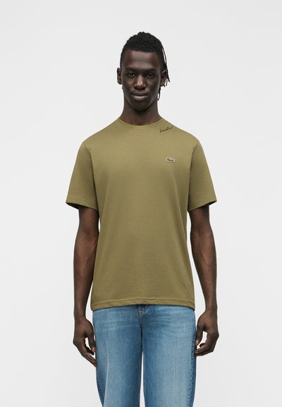 Млад мъж със тъмна кожа и плитки, облечен в маслиненозелен тениска Lacoste и сини дънки, стоящ срещу светъл едноцветен фон.