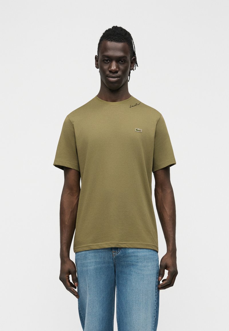 Νεαρός άνδρας με σκούρο δέρμα και πλεγμένα μαλλιά, φορώντας ελιάς πράσινη μπλούζα Lacoste και μπλε τζιν, στέκεται μπροστά σε ανοιχτόχρωμο φόντο.