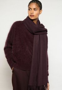 Plūmju šalle, izgatavota no maiga, teksturēta auduma ar frēžotām malām, brīvi apvīta ap kaklu, valkāta kopā ar atbilstošu pūkainu džemperi.