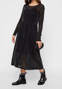 Un vestido negro de manga larga con una superposición brillante y transparente, un torso ajustado y una falda fluida; combinado con botas negras hasta el tobillo.