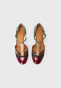 Multi-colored sandals featuring a green leather upper, braided beige accent, and burgundy patent toe cap, with a t-strap design and adjustable buckle.