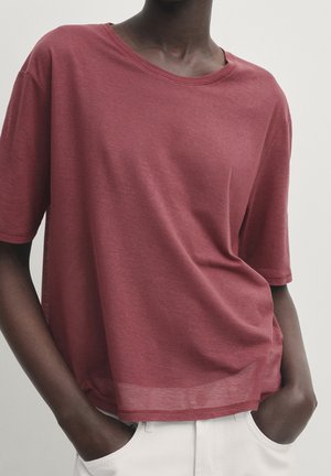 Personne à la peau foncée portant un t-shirt bordeaux ample et un pantalon blanc, les mains dans les poches sur un fond uni.