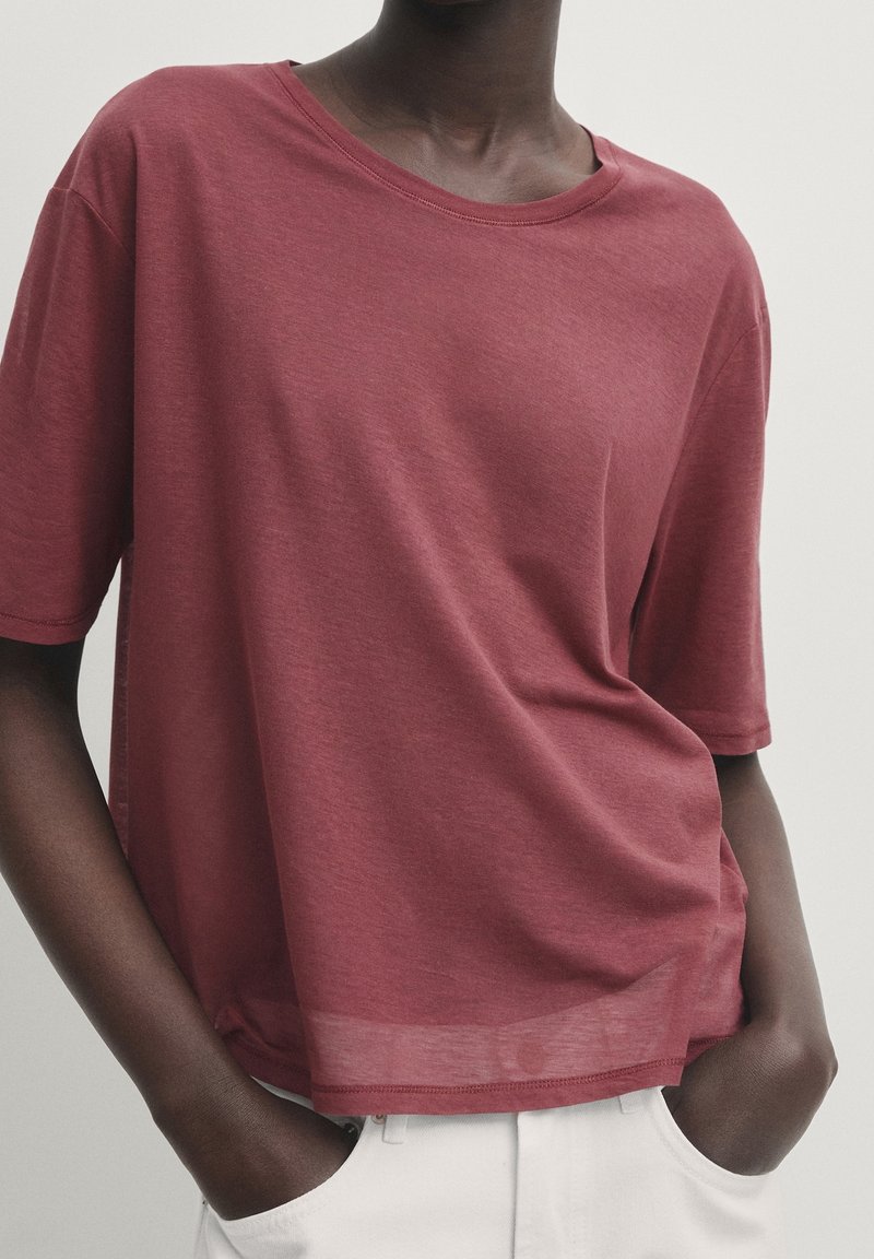 Personne à la peau foncée portant un t-shirt bordeaux ample et un pantalon blanc, les mains dans les poches sur un fond uni.
