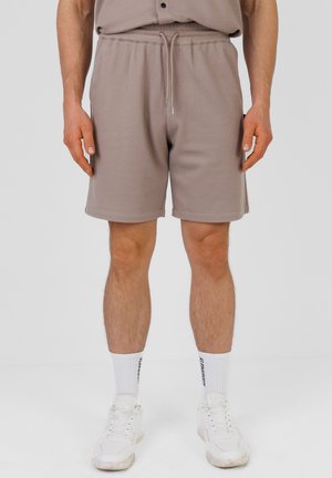 Man draagt taupekleurige korte broek met trekkoord, witte sportsokken met zwarte tekst en witte sneakers, staand tegen een effen achtergrond.