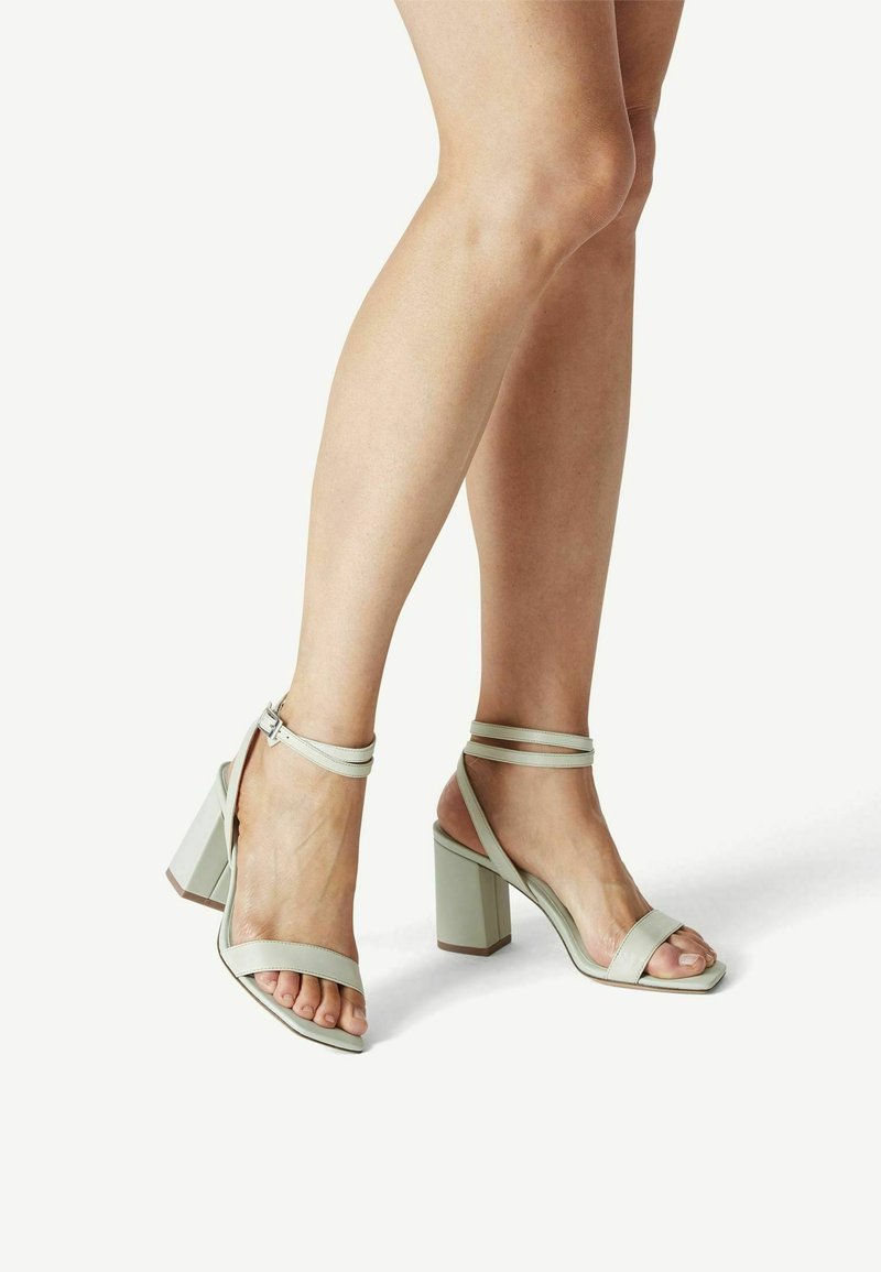 Jambes portant des sandales à talons hauts gris clair avec des brides à la cheville, debout sur un fond blanc.