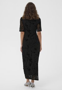 Čierne kvietkované sametové šaty s priesvitnými krátkymi rukávmi a priliehavým strihom, siahajúcim tesne nad členky, v kombinácii s svetlosivou obuvou.