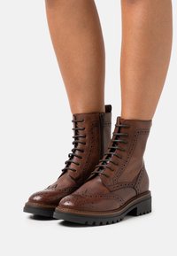 Bottes cheville en cuir marron avec un design brogue, comprenant des lacets, une fermeture éclair latérale et une semelle robuste pour une bonne adhérence.