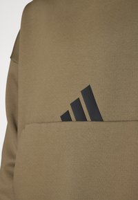Brūns džemperis ar priekšējo kabatu un melnu trīsu joslu logo. Tekstūra šķiet gluda, un audumam, visticamāk, ir mīksts džemperu materiāls.