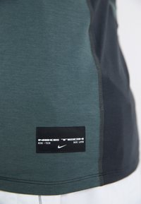 Camisa base de camada da Nike em verde escuro e preto. Apresenta uma textura suave, design contrastante e um emblema com texto branco.