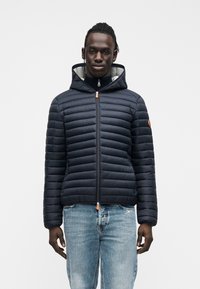 Tengerészkék steppelt pehelydzseki vízszintes csíkmintával, kapucnival, elülső cipzárral, narancssárga cipzárhúzóval és szürke belső béléssel. Világoskék farmernadrággal viselve.