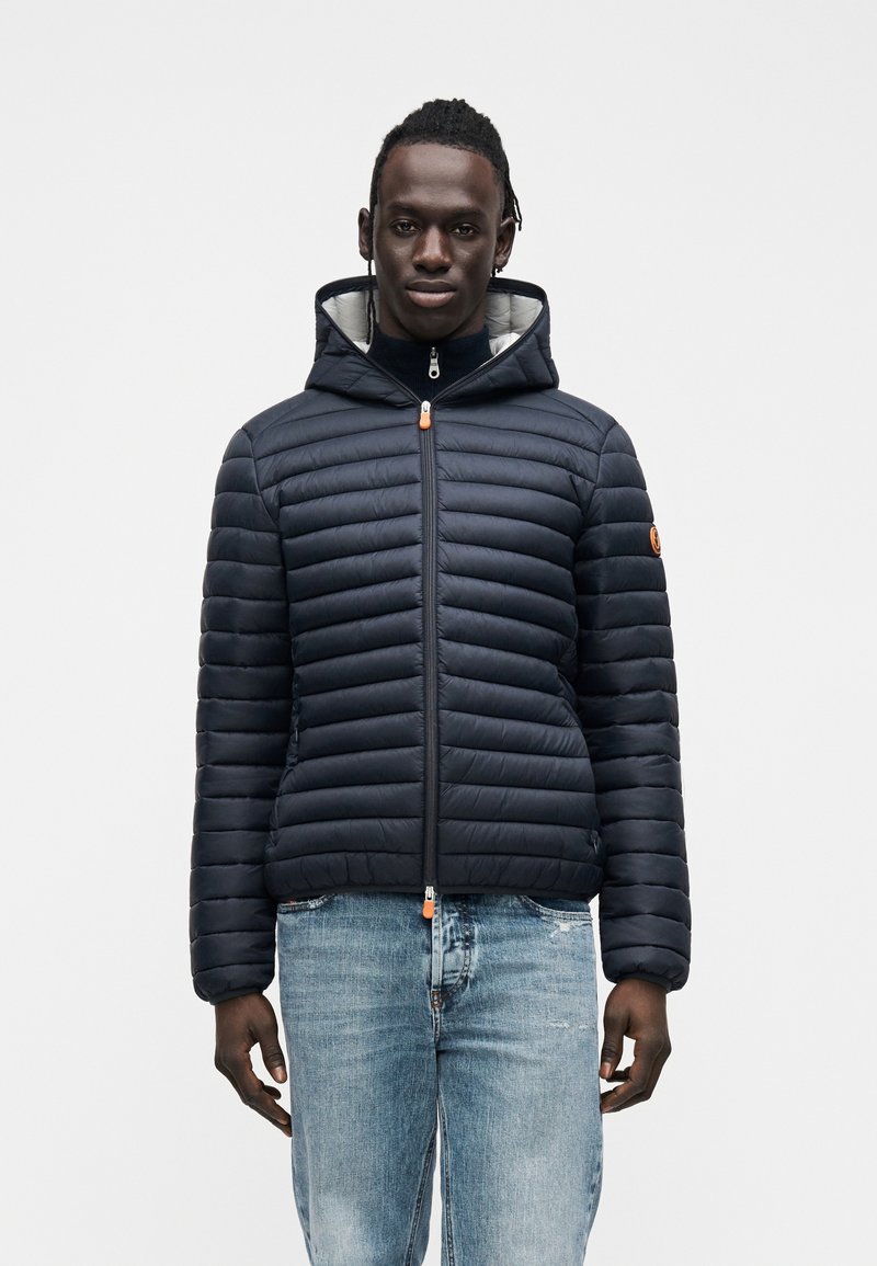 Tengerészkék steppelt pehelydzseki vízszintes csíkmintával, kapucnival, elülső cipzárral, narancssárga cipzárhúzóval és szürke belső béléssel. Világoskék farmernadrággal viselve.