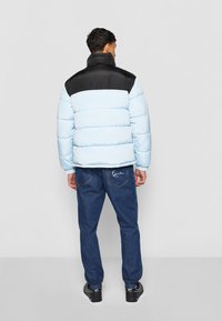 Lys blå pufferjakke med en sort overdel, kviltet design, høy krage, kombinert med mørkeblå jeans og svarte joggesko.