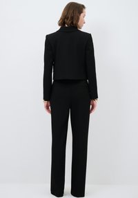 Completo nero sartoriale con giacca corta e pantaloni dritti. Il tessuto appare liscio, con un classico risvolto e discreti dettagli delle cuciture.