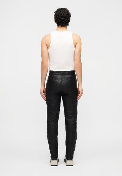 Άτομο με σγουρά σκουρόχρωμα μαλλιά, ντυμένο με λευκό αμάνικο τοπ, μαύρο δερμάτινο παντελόνι ψηλής μέσης και μαύρα παπούτσια με λευκά τακούνια σε σχήμα μπλοκ, όρθιο.