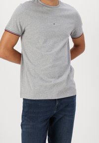 Harmaa puuvillapaita, jossa on pyöreä pääntie ja lyhyet hihat, punaisilla ja laivastonsinisen raidalla varustettuna, sekä pieni Tommy Hilfiger -logo rinnassa.