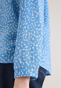 Gros plan sur une personne portant une chemise à manches longues bleue avec des motifs abstraits blancs et un jean en denim bleu foncé.