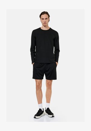 Man standing with hands in pockets wearing black long-sleeve shirt, black shorts, white socks, and black-and-white sneakers on a white background.