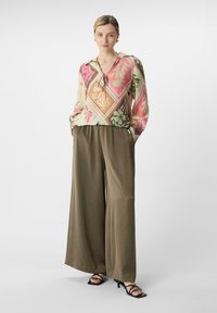 Bunt bedruckte Bluse mit Blumenmuster, V-Ausschnitt und Bändern, kombiniert mit hochgeschnittenen, weit geschnittenen, olivgrünen Hosen und schwarzen, geschlossenen High-Heels.