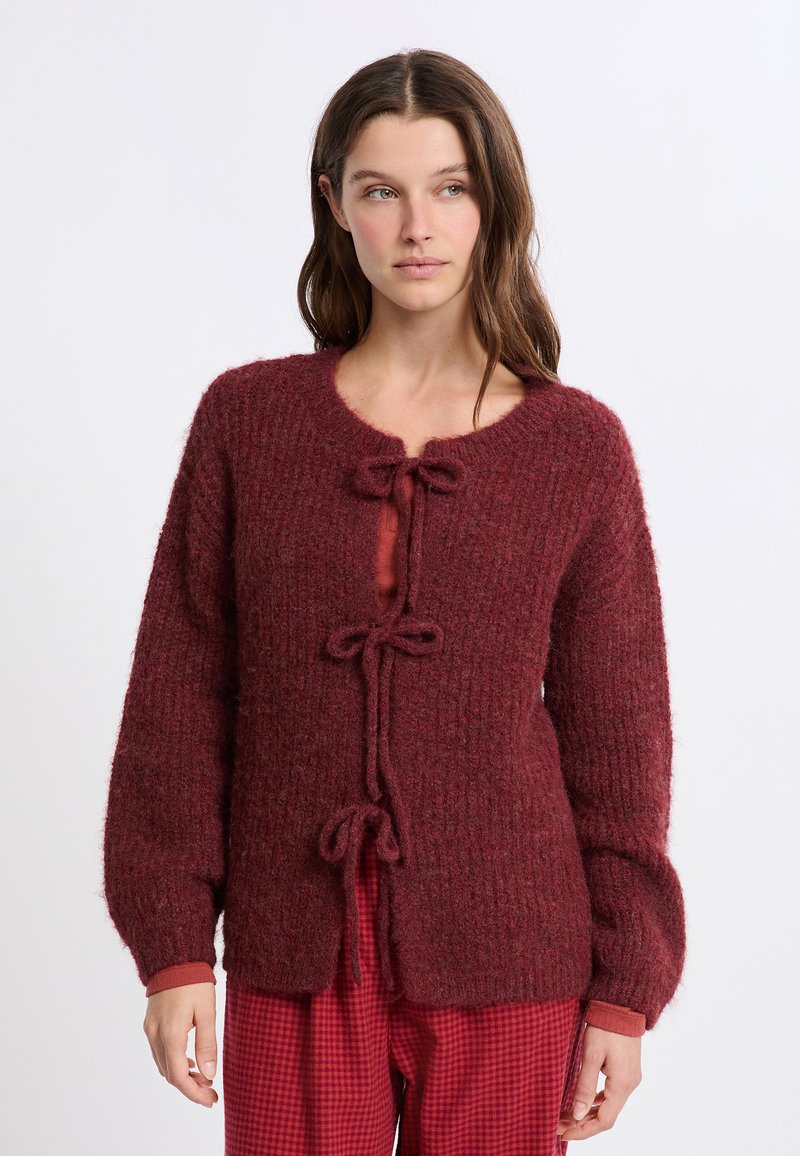 Cardigan in borgogna a maglia con finitura testurizzata, scollatura rotonda e fiocchi lungo il davanti. Maniche lunghe con polsini a costine.