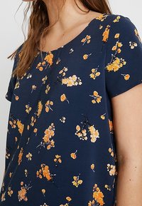 Tummansininen kukallinen paita, jossa on hajautettuja oransseja ja keltaisia kukkakuoseja, lyhyet hihat ja pyöreä kaula-aukko pehmeässä kankaassa.