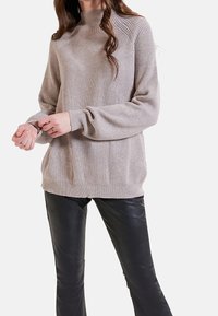 Světle béžový žebrovaný svetr s vysokým límcem, má dlouhé rukávy a manžety. Kombinován s černými koženými kalhotami pro kontrast.