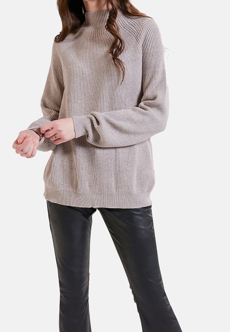 Světle béžový žebrovaný svetr s vysokým límcem, má dlouhé rukávy a manžety. Kombinován s černými koženými kalhotami pro kontrast.