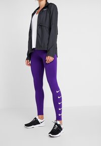 Musta kevyt takki, valkoinen tanktoppi, violetit napakat legginsit, joissa on valkoiset Nike-logot, ja mustat urheilukengät valkoisilla yksityiskohdilla.