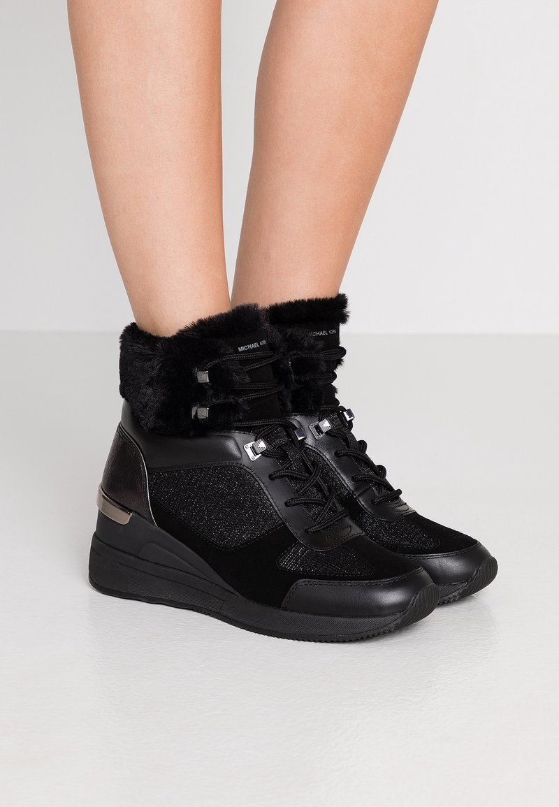 Personne portant des bottines noires à talon compensé avec bordure en fausse fourrure, accents métalliques et laçage à l'avant, debout sur un sol blanc brillant.