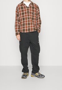 Brun och vit rutigt flanellskjorta, svarta cargobyxor med sidofickor, grå och gula sportskor, stående pose.