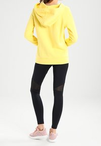 Gul hoodie, svarta leggings med mesh-accenter och rosa träningsskor. Släta tyger, avslappnad passform och kontrasterande detaljer.