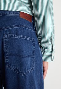 Jeans iz jeansa z zadnjim žepom in rjavim usnjenim oznako blagovne znamke. Imajo temno modro tkanino in opazno šivano vzorčenje. Delno vidna je tudi rokav črtaste zelene srajce.