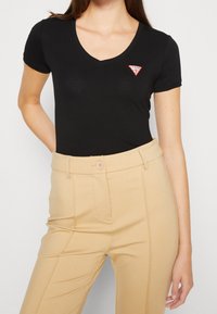 Zwarte katoenen t-shirt met een V-hals en korte mouwen. Heeft een rood driehoeklogo op de linkerborst. Gecombineerd met beige high-waisted broeken.
