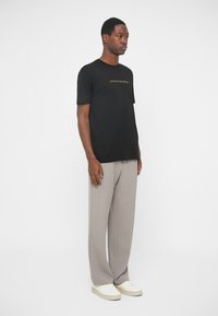 Άνδρας με μαύρο μπλουζάκι Armani Exchange, μπεζ παντελόνι φάρδους και λευκά αθλητικά παπούτσια, που στέκεται μπροστά σε λευκό φόντο.