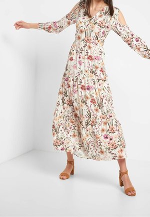 Kvinde iført en lang blomsterprintet kjole med cut-outs ved skuldrene og tanfarvede ankelremssandaler med blokhæle, poserer med armene udstrakte.